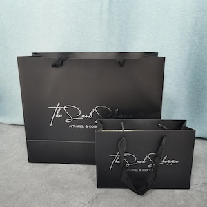 May include: Two black shopping bags of different sizes with black rope handles. The bags have the text "The Snob Shoppe" in a cursive font, with "Apparel & Cosmetics" below.