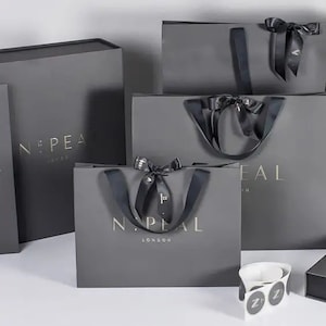 May include: A collection of gray gift bags and boxes with gold lettering. The bags have black ribbon handles and bows. The text "N:PEAL LONDON" is visible on the bags. A few white stickers with a black "Z" are also visible.