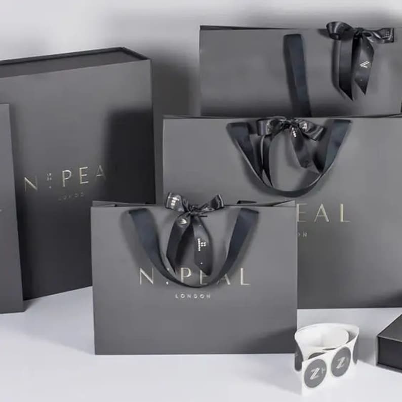 May include: A collection of gray gift bags and boxes with gold lettering. The bags have black ribbon handles and bows. The text "N:PEAL LONDON" is visible on the bags. A few white stickers with a black "Z" are also visible.