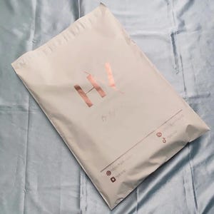 May include: A light beige shipping package with a rose gold logo and text. The logo features the letters "HV" in a stylized design. Additional text includes a website address and social media handles.