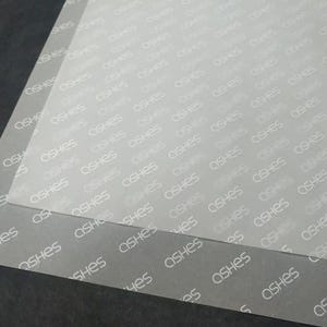 Puede incluir: Una hoja rectangular de material semitransparente y esmerilado con un patrón repetitivo de la palabra ASHES impresa en una fuente blanca sans-serif. La superficie tiene un acabado mate y está colocada sobre una base de color oscuro.