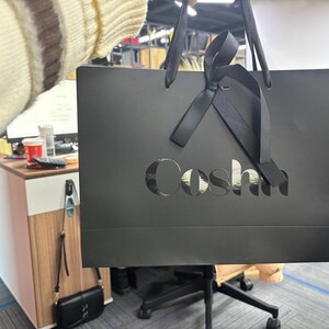 May include: A black shopping bag with the word "Coshi" cut out on the front. The bag has black ribbon handles and a bow. The background shows an office setting with a desk and chair.