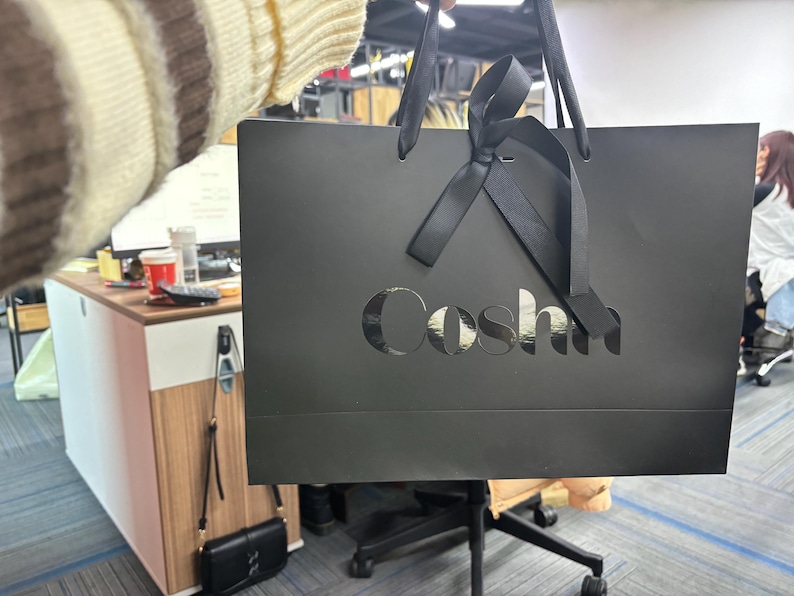 May include: A black shopping bag with the word "Coshi" cut out on the front. The bag has black ribbon handles and a bow. The background shows an office setting with a desk and chair.