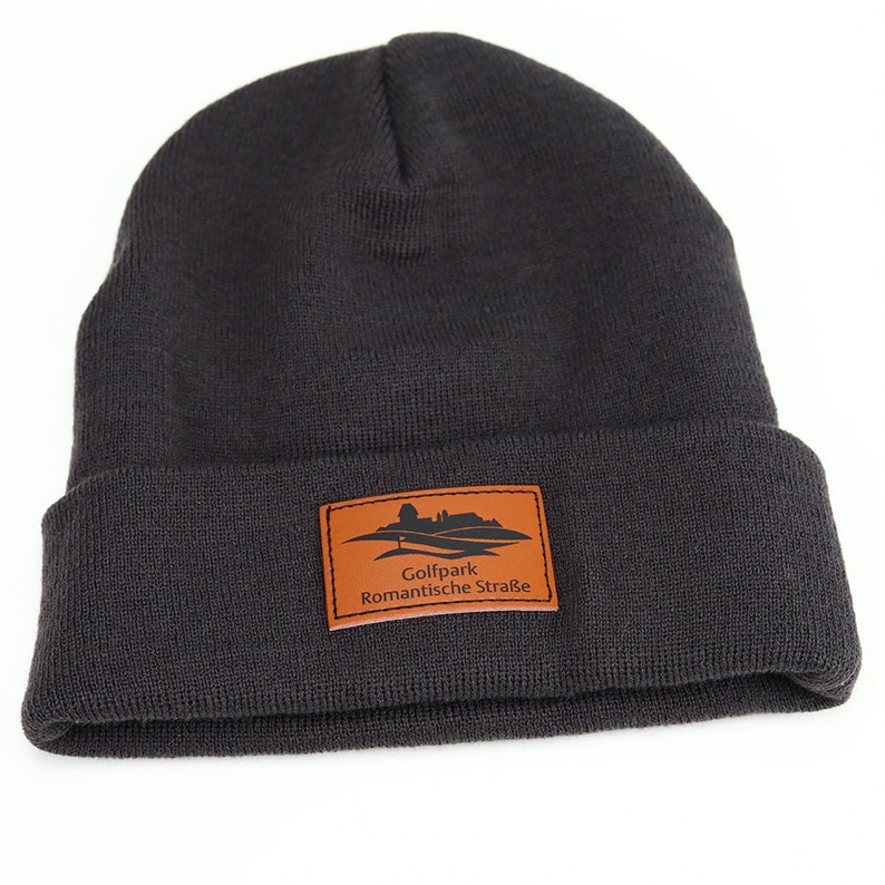 May include: A dark gray knit beanie with a brown leather patch that says "Golfpark Romantische Stra&szlig;e".