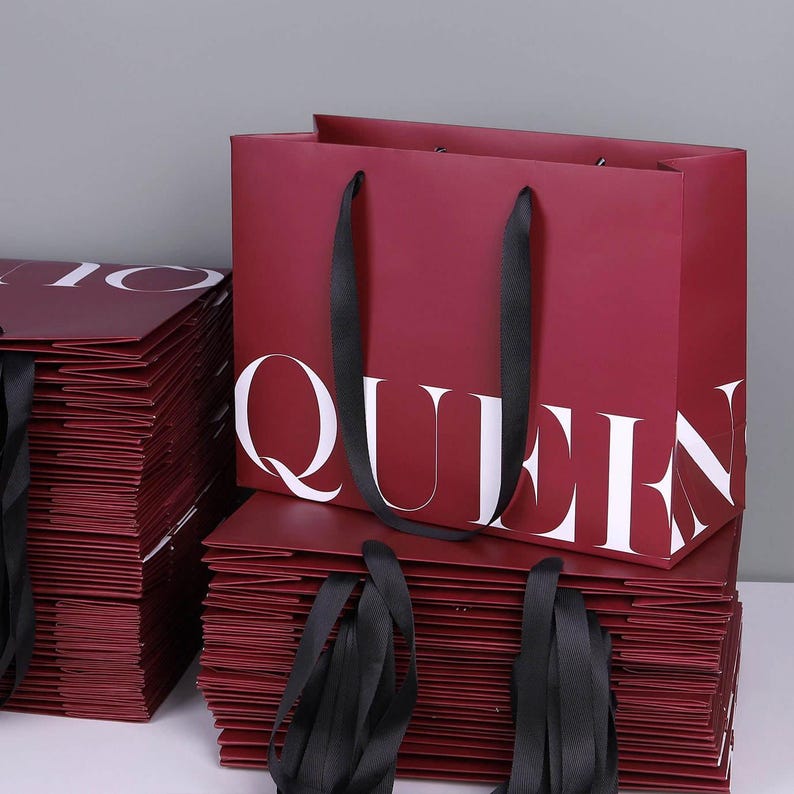 May include: Burgundy shopping bags with black ribbon handles are stacked. The word "QUEEN" is printed in large white letters on the front of the bag. Additional bags with the word "IO" are visible.