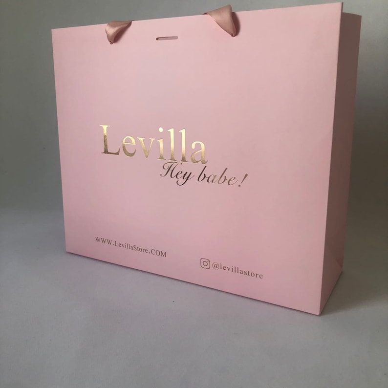 May include: A pink shopping bag with gold lettering that reads "Levilla Hey babe!" and the website address. The bag has pink ribbon handles.