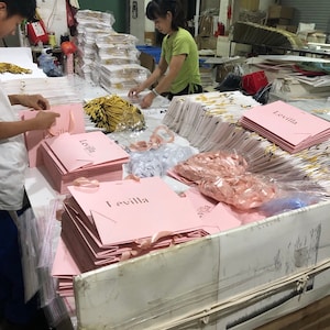 May include: Workers assembling pink Levilla shopping bags with ribbon handles. The bags are stacked on a table, with additional bags and materials in the background. The bags have the word "Levilla" printed on them.