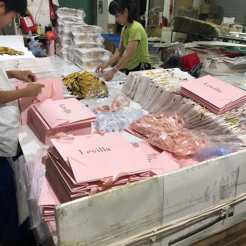 May include: Workers assembling pink Levilla shopping bags with ribbon handles. The bags are stacked on a table, with additional bags and materials in the background. The bags have the word "Levilla" printed on them.