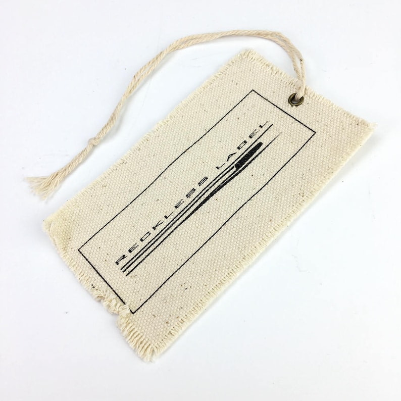 May include: A white cotton fabric tag with frayed edges and a black string attached. The tag has a black rectangle with the text "RECKLESS LABEL" printed inside.