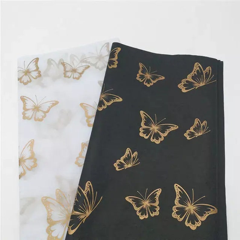 K&ouml;nnte beinhalten: Zwei Seidenpapiere, eines wei&szlig; und eines schwarz, werden gezeigt. Beide Bl&auml;tter sind mit goldenen Schmetterlingsdesigns verziert. Die Schmetterlinge sind unterschiedlich gro&szlig; und &uuml;ber das Papier verstreut, wodurch ein zartes und elegantes Muster entsteht.