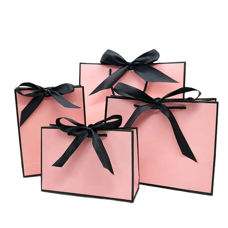 May include: Four pink gift bags of varying sizes, each adorned with a black satin bow. The bags have black trim and matching black handles. The bags are arranged against a white background.