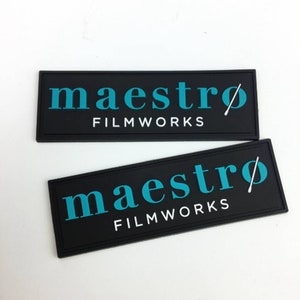 May include: Two black rectangular patches with white text that reads "maestro FILMWORKS" and a stylized white conductor's baton. The text is in a teal blue color.