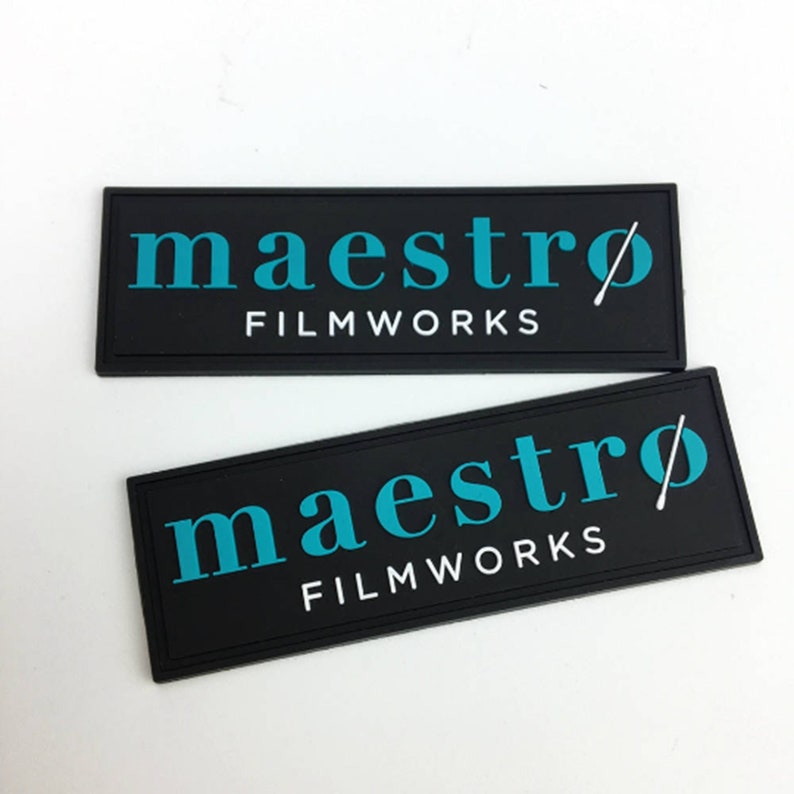 May include: Two black rectangular patches with white text that reads "maestro FILMWORKS" and a stylized white conductor's baton. The text is in a teal blue color.