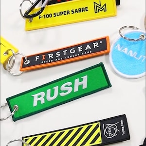May include: Collection of embroidered keychains in various colors, including red, green, yellow, and black. The keychains feature text such as "HIDE MIC", "Right On Time Delivery Services", "F-100 SUPER SABRE", and "RUSH".