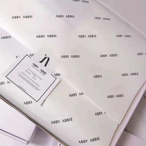 Puede incluir: Papel de seda blanco con el texto negro repetido ABBY ABBIE, acompañado de una tarjeta rectangular blanca con un logotipo negro y el texto ABBY ABBIE, que incluye un mensaje impreso agradeciendo al cliente por su compra.