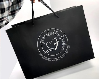 May include: Black paper shopping bag with a white logo that says "gracefully broken" with a heart and cross design. The text below the logo says "Be Blessed. Be Beautiful. Be You."
