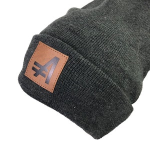 May include: A dark gray beanie with a brown leather patch featuring a black stylized letter "A" with a diagonal line through it.