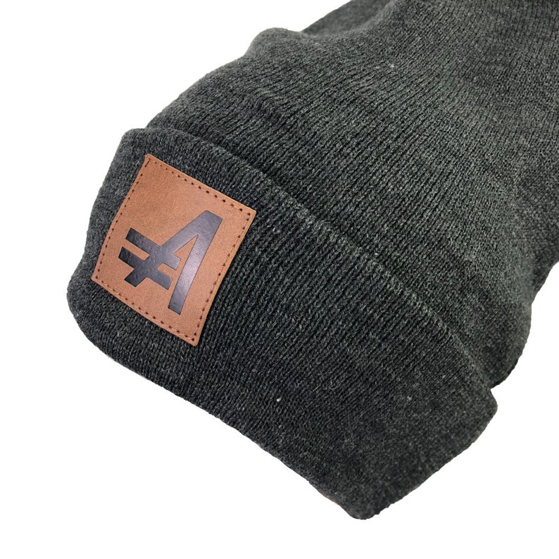 May include: A dark gray beanie with a brown leather patch featuring a black stylized letter "A" with a diagonal line through it.