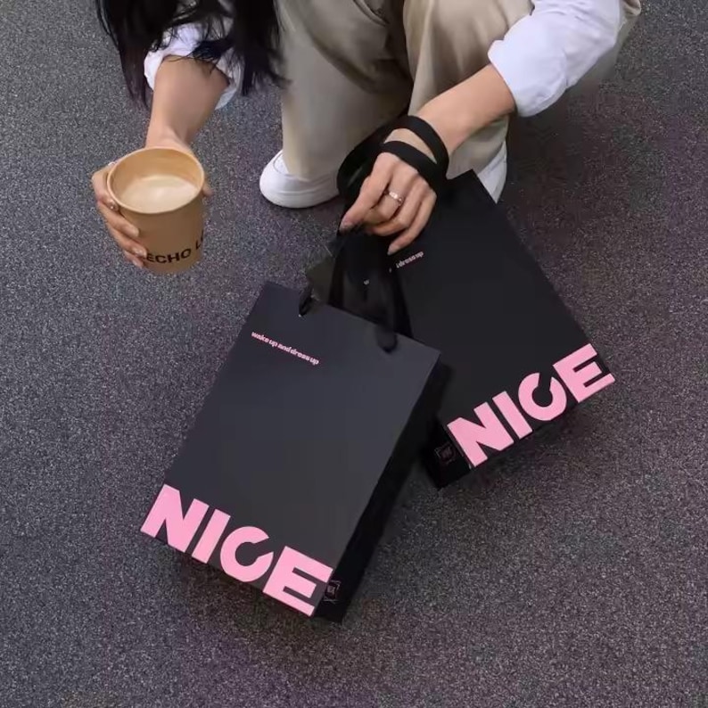 May include: Two black shopping bags with the word "NICE" in pink block letters. One bag has the text "wake up and dress up" in pink. A person is holding the bags and a coffee cup.