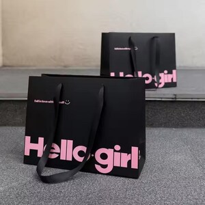 May include: Two black shopping bags with pink text that reads "Hello girl." One bag has the phrase "fall in love with self" and a smiling face. The bags have black handles and are on a gray surface.