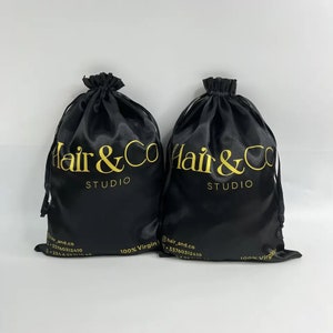 May include: Two black satin drawstring bags. Gold text on the bags reads "Hair & Co STUDIO". The bags are labeled "100% Virgin". Contact details are printed at the bottom of each bag.