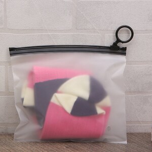 Pu&ograve; includere: Una bustina trasparente in plastica con cerniera e tiretto ad anello nero. La bustina contiene un paio di calzini a righe rosa, bianche e nere.