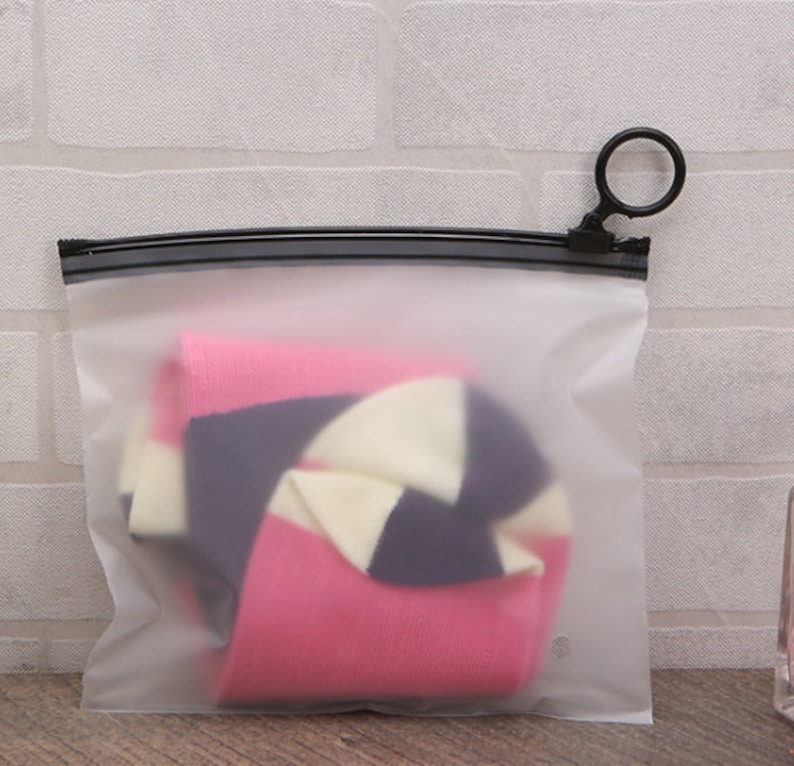 Pu&ograve; includere: Una bustina trasparente in plastica con cerniera e tiretto ad anello nero. La bustina contiene un paio di calzini a righe rosa, bianche e nere.