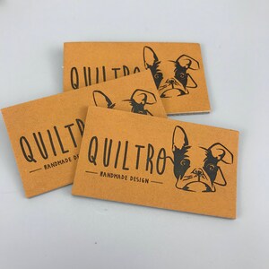 Op de afbeelding: Drie bruine papieren etiketten met zwarte tekst en een zwart silhouet van een Franse bulldog. De tekst op de etiketten luidt "Quiltro - Handmade Design".