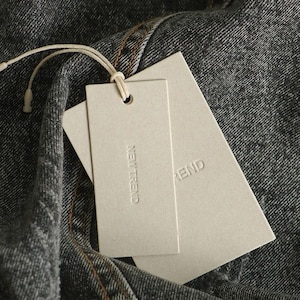 May include: Two rectangular off-white paper tags attached with a thin beige string to a piece of dark gray denim fabric. The top tag features the embossed text NEW TREND, while the bottom tag partially shows the same embossed text.