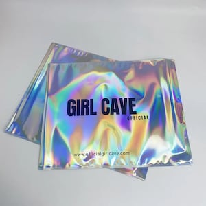 Puede incluir: Dos sobres de env&iacute;o plateados iridiscentes con el texto "GIRL CAVE OFFICIAL" y el sitio web "www.officialgirlcave.com" impresos en ellos.