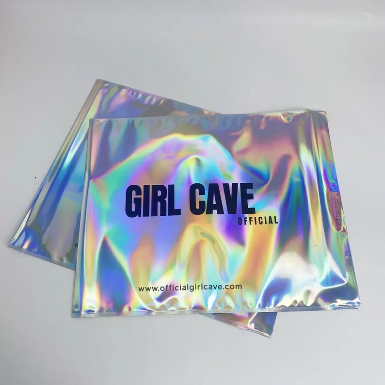 Puede incluir: Dos sobres de env&iacute;o plateados iridiscentes con el texto "GIRL CAVE OFFICIAL" y el sitio web "www.officialgirlcave.com" impresos en ellos.