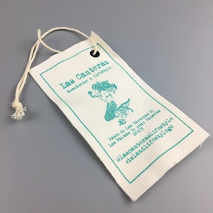 May include: A white fabric tag with a teal blue border and text. The tag features a mermaid illustration and the text "Las Canteras Beachwear & Lifestyle." The text also includes the address "Paseo de Las Canteras 20, Las Palmas de Gran Canaria, SPAIN" and the social media handles "@lascanteraslifestyle #islandlifestylega".