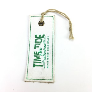 May include: White fabric tag with a green embroidered logo that says "Time and Tide Collection" and "Registered Trademark". The tag has a brown metal eyelet and a brown string attached.