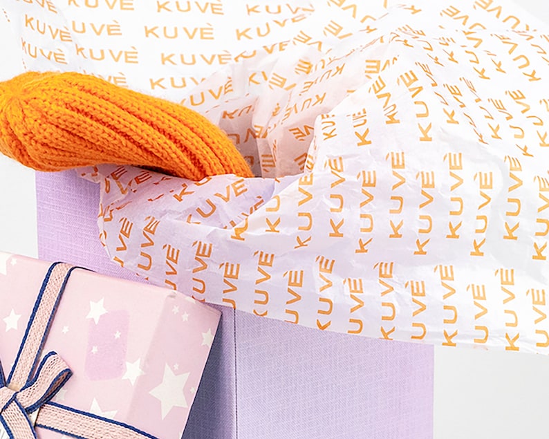 K&ouml;nnte beinhalten: Eine hellviolette Geschenkbox mit orangefarbenem Seidenpapier, auf dem das Wort "KUVE" steht. Ein orangefarbener Strickartikel befindet sich in der Box. Ein rosa Geschenk, verpackt mit einem Sternmuster und marineblauem Band, ist im Vordergrund.