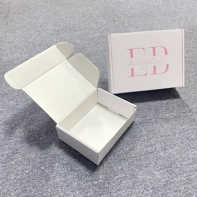 May include: White cardboard box with a fold-over flap. The box is open and the inside is visible. The box has the text "Beauty Essentials" and the letters "ED" in pink.