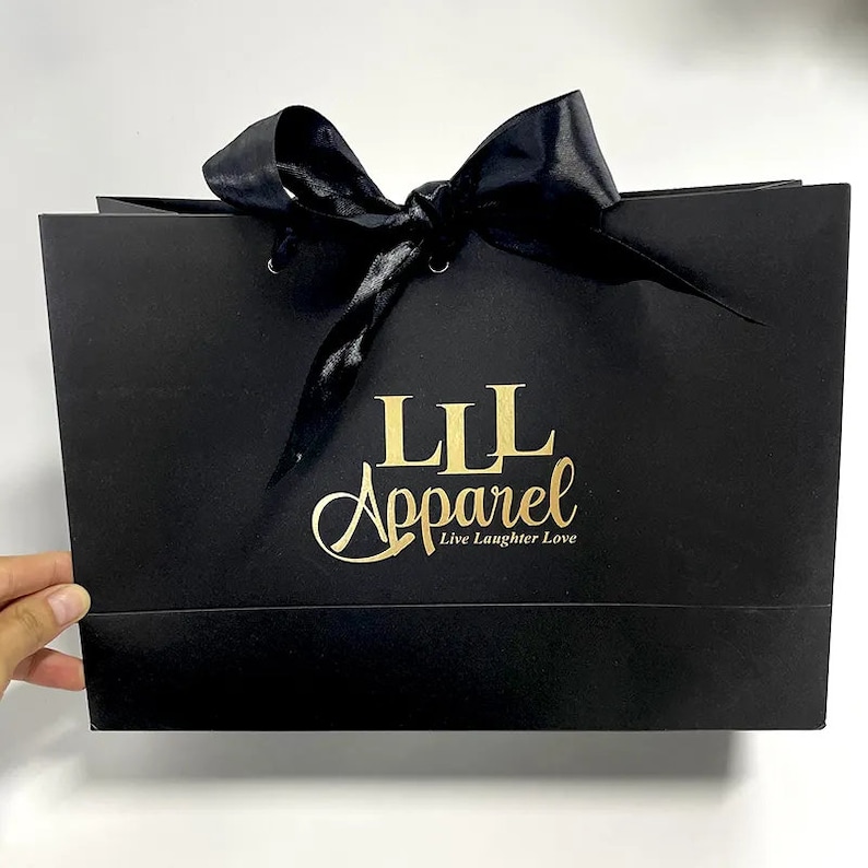 May include: Black gift bag with a shiny black ribbon tied in a bow. The bag has the gold text "LLL Apparel" and the tagline "Live Laughter Love". The bag is rectangular and has a glossy finish.