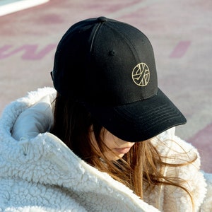 以下が含まれることがあります： 金の刺繍が施された円形のロゴが付いた黒いベースボールキャップ。ロゴは円の中にスタイリッシュなXが描かれています。