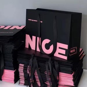 May include: Black shopping bags with pink text that reads "NICE" and "wake up and dress up". The bags have black ribbon handles and are stacked on top of each other. The edges of the bags are pink.