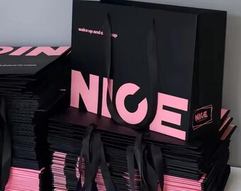 May include: Black shopping bags with pink text that reads "NICE" and "wake up and dress up". The bags have black ribbon handles and are stacked on top of each other. The edges of the bags are pink.