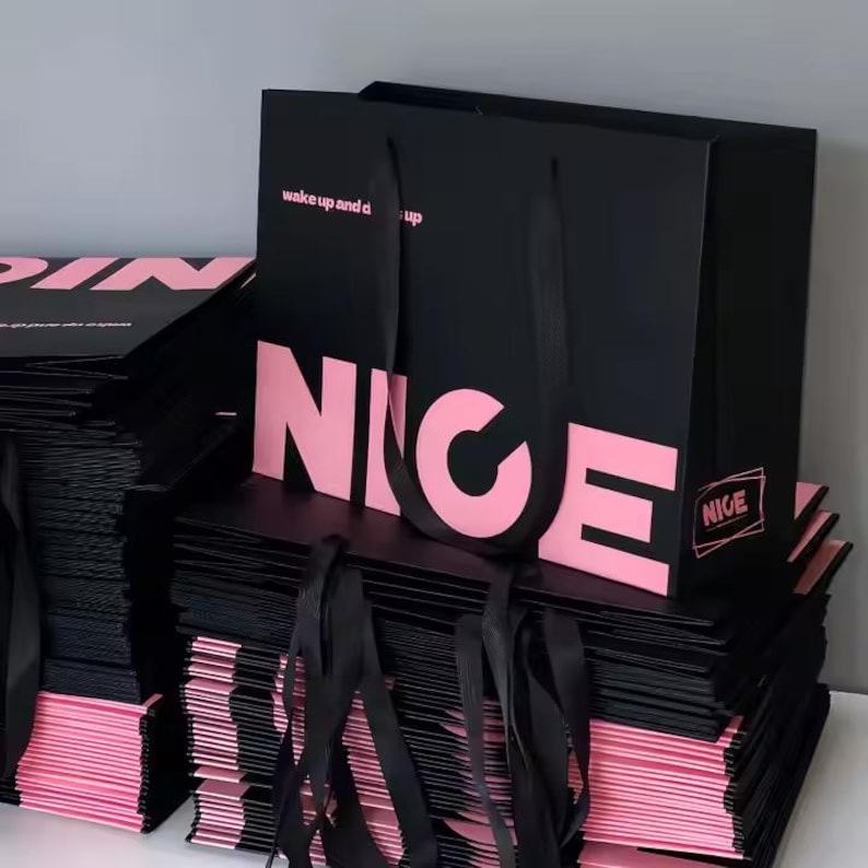 May include: Black shopping bags with pink text that reads "NICE" and "wake up and dress up". The bags have black ribbon handles and are stacked on top of each other. The edges of the bags are pink.