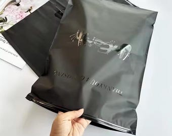 May include: Two black poly mailer shipping bags. One bag is in the foreground, held by a hand. The bags have a silver design and the words "THANK YOU 4 CHOOSING" printed on them.