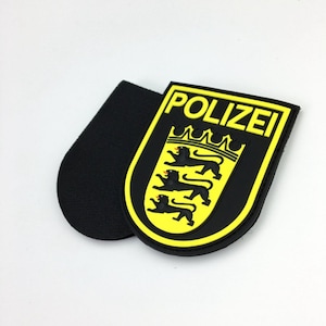 May include: A black and yellow rubber patch with the word "POLIZEI" in yellow letters and a black coat of arms with three lions. The patch has a black hook and loop backing.