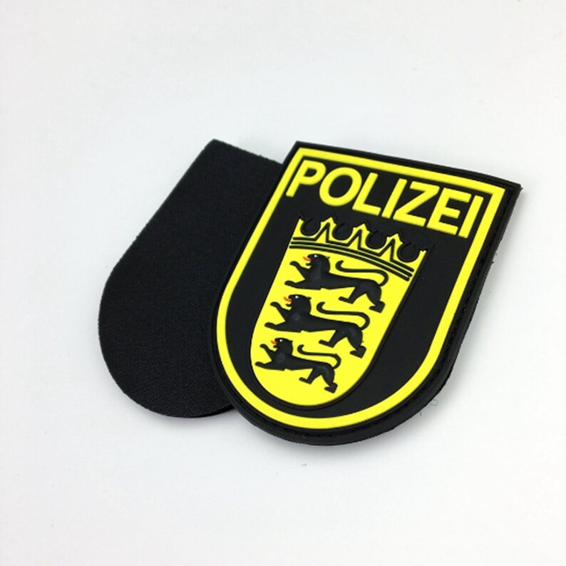 May include: A black and yellow rubber patch with the word "POLIZEI" in yellow letters and a black coat of arms with three lions. The patch has a black hook and loop backing.