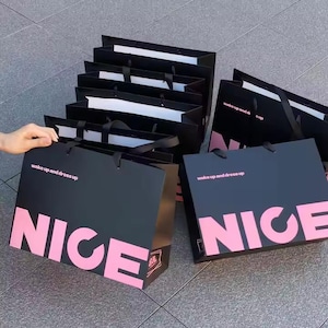 May include: A collection of black shopping bags with the word "NICE" in large pink letters. The bags also have the text "wake up and dress up" in pink. The bags have black handles.