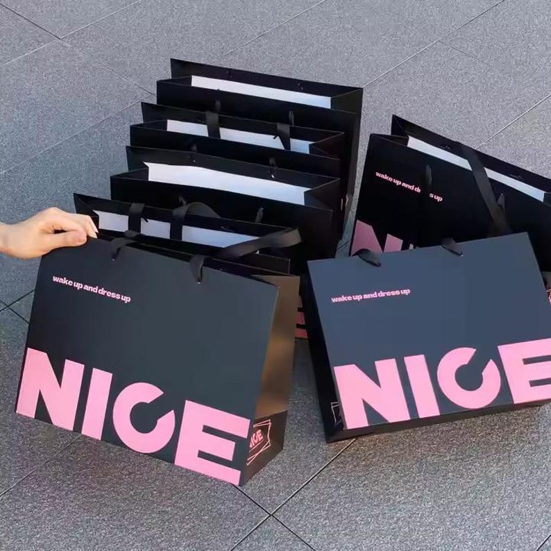May include: A collection of black shopping bags with the word "NICE" in large pink letters. The bags also have the text "wake up and dress up" in pink. The bags have black handles.