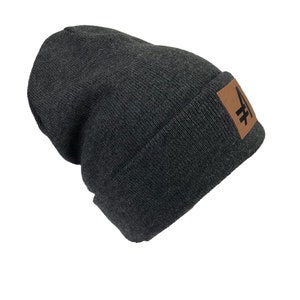 May include: A dark gray beanie with a brown leather patch featuring a black graphic of a stylized letter "A".