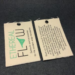 May include: Two white fabric tags with black text and a green logo. The text on the tags reads "Ethereal Flow Swimsuits and accessories for the modern, edgy trendsetter." and "Thank you for trusting EF to enhance your swim/beach wardrobe. We take pride in the look and quality of our products and hope you will be greatly satisfied with your purchase. Thank you for shopping with us and we appreciate your business! Like us on Facebook. Follow & tag us on Instagram @etherealflowswin and #EFSwim in one of our pieces for your chance to be featured. www.etherealflowswim.com"