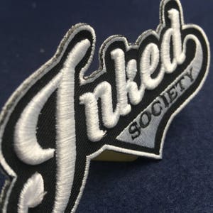 Puede incluir: Parche bordado blanco con el texto "Inked Society" en blanco y negro. El parche tiene la forma de un logotipo de béisbol.