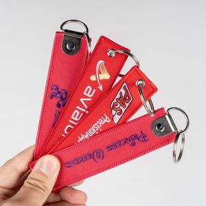 May include: A hand holding a fan of red fabric keychains. Each keychain has a silver ring and features embroidered text and logos in white and purple. The keychains are for aviation, precision, and other brands.