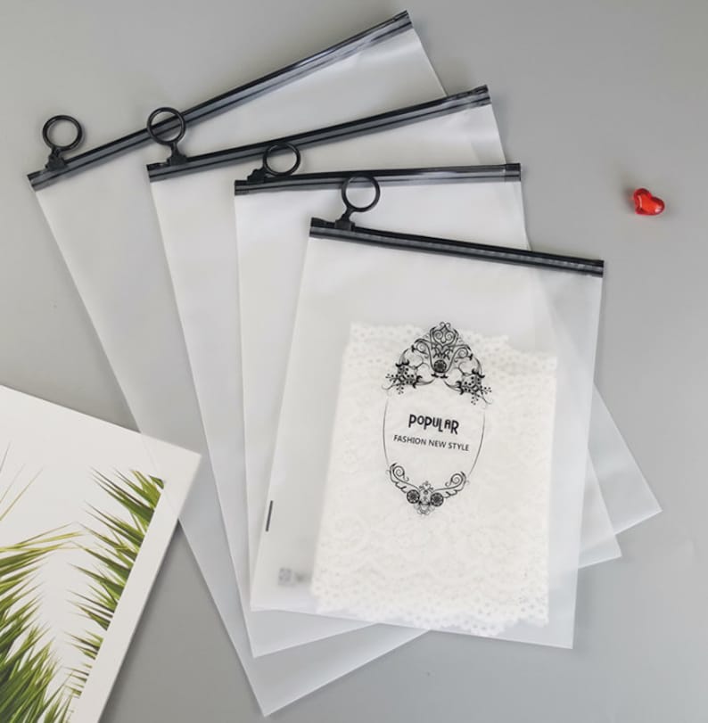 Pu&ograve; includere: Quattro buste di plastica trasparente con cerniera, con cerniere nere e bordi neri. Le buste sono impilate l'una sull'altra. La busta superiore presenta un design in bianco e nero con il testo "POPULAR FASHION NEW STYLE".